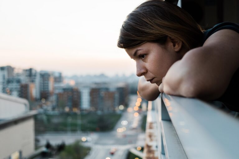 A woman is standing on her balcony, leaning with her arms folded on the railing in front of her. She looks out to the city below with a somber expression.